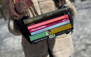 A person wearing black gloves is holding a stack of books in front of their torso. The books are Held by Anne Michaels, The Sleeping Car Porter by Suzette Mayr, How to Pronounce Knife by Souvankham Thammavongsa, Hellgoing by Lynn Coady, The Sentimentalists by Johanna Skibsrud, Late Nights on Air by Elizabeth Hay and A Good House by Bonnie Burnard.