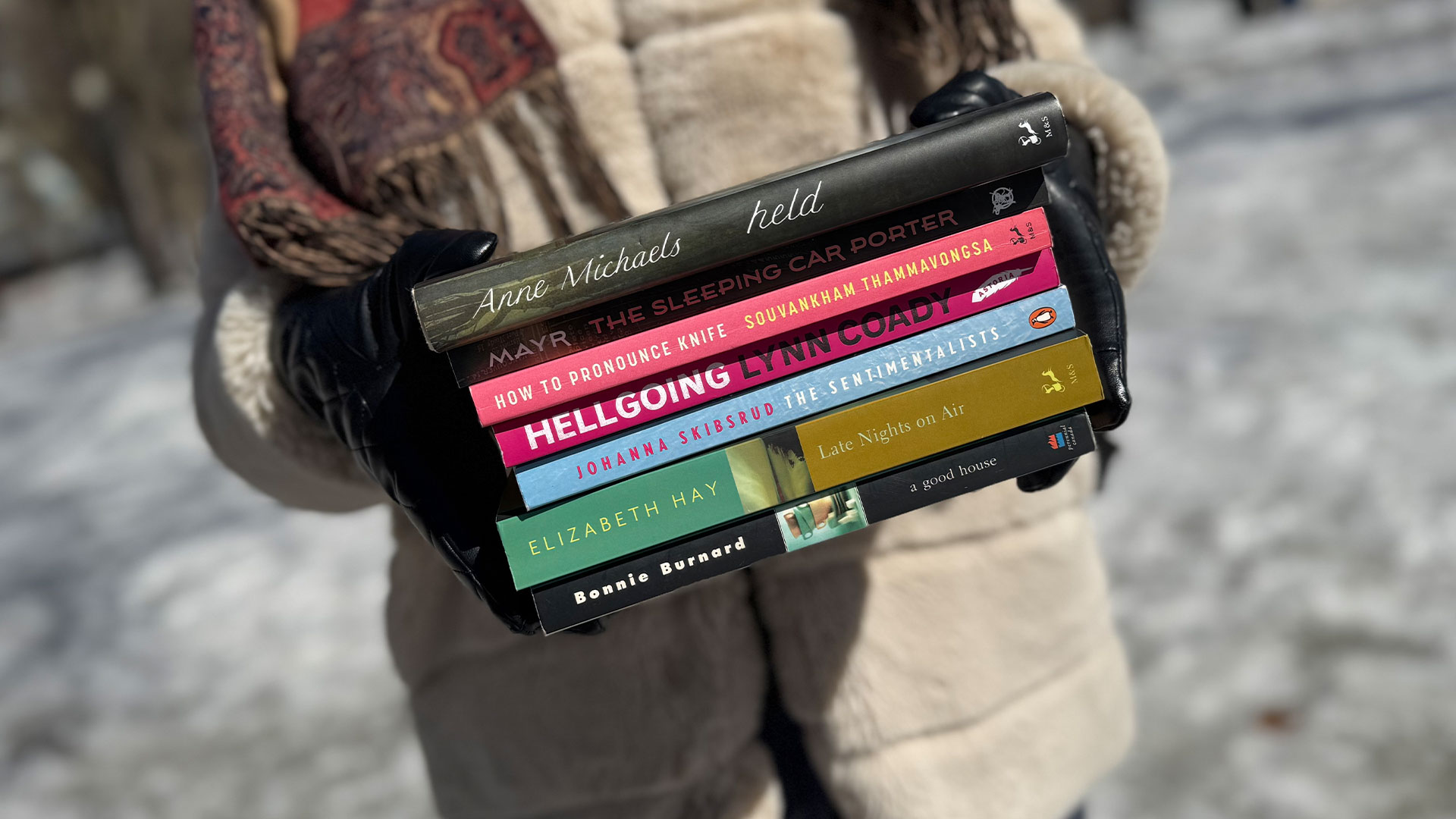 A person wearing black gloves is holding a stack of books in front of their torso. The books are Held by Anne Michaels, The Sleeping Car Porter by Suzette Mayr, How to Pronounce Knife by Souvankham Thammavongsa, Hellgoing by Lynn Coady, The Sentimentalists by Johanna Skibsrud, Late Nights on Air by Elizabeth Hay and A Good House by Bonnie Burnard.